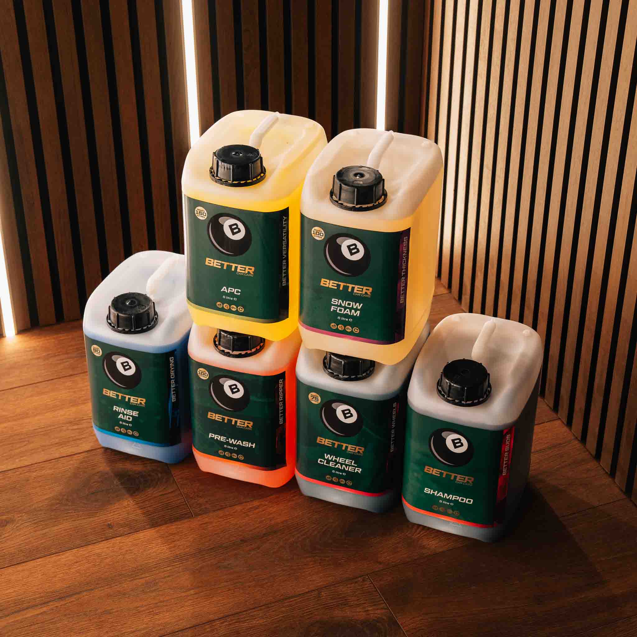 Six Better brand cleaning product jugs arranged on a wooden floor against a slatted wood wall. The set includes Blue, White, Orange, and Red variants of shampoo, prewash, snow foam, and shower foam.