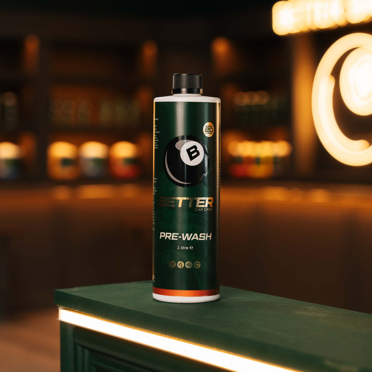 A bottle of Better Car Care Pre-Wash stands on a dark green counter in a dimly lit, warm-toned bar or lounge setting. The bottle is dark green with a black cap and features the Better Car Care logo and branding.