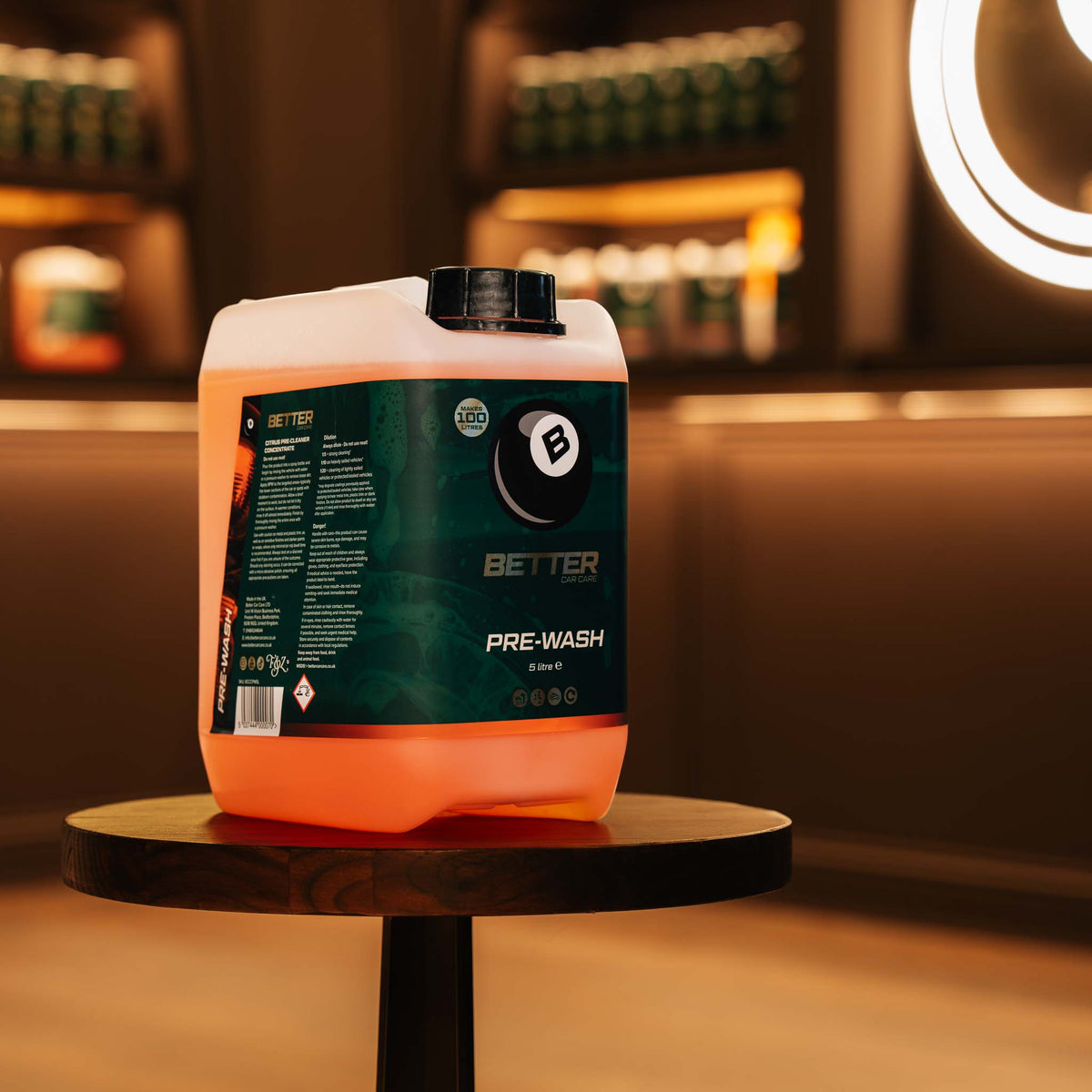 A large translucent orange and white plastic jerrycan of Better Pre-Wash liquid cleaner sits on a round wooden table in a warmly lit indoor setting with blurred shelves and lighting in the background.