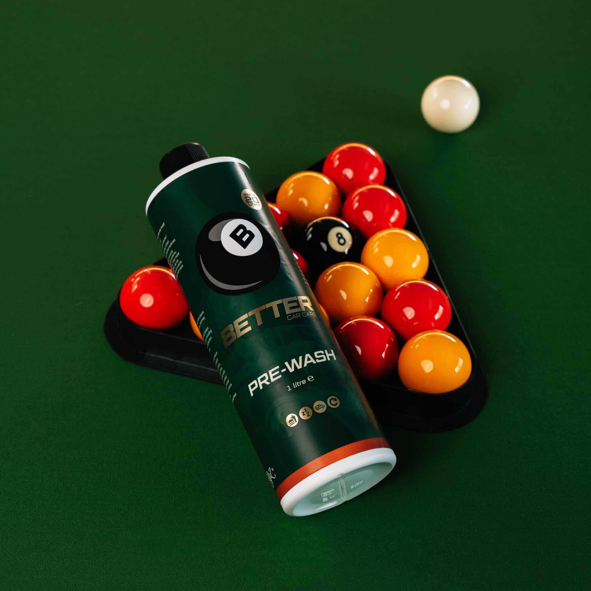 A bottle of Better Pool Pre-Wash liquid lies on a green pool table next to a rack of colorful billiard balls, with one white ball in the background.