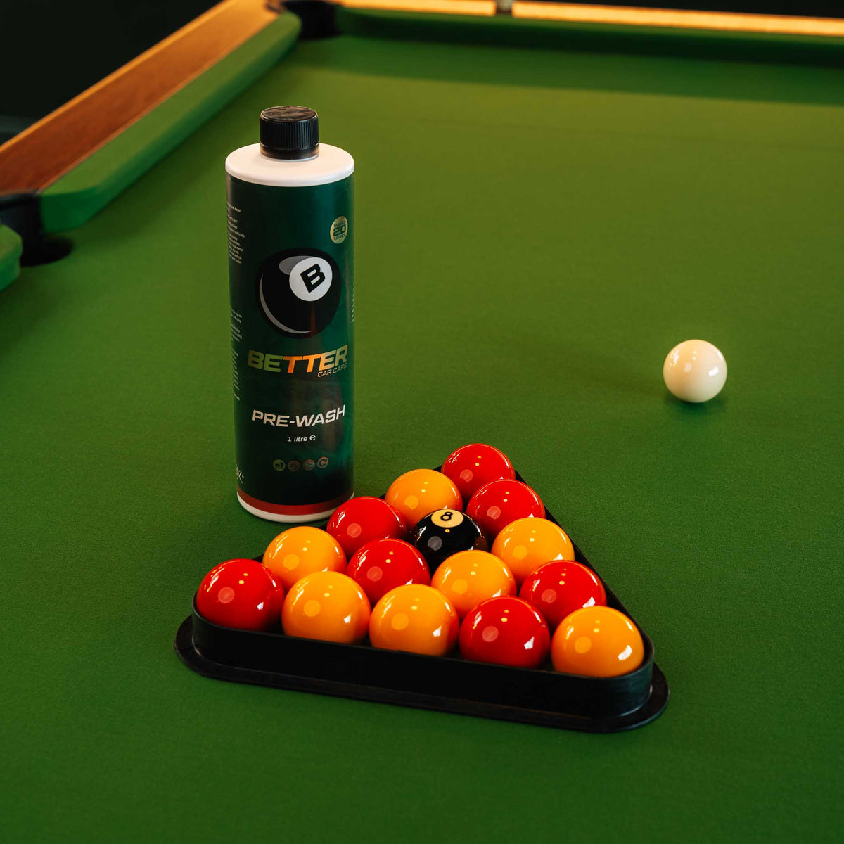 A bottle of Better Pre-Wash pool ball cleaner stands on a green pool table next to a triangle rack of colored pool balls, with a white cue ball nearby.