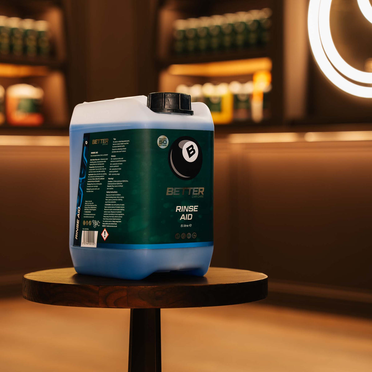 A large plastic canister of Better Brand Rinse Aid sits on a small round wooden table in a warmly lit, blurred indoor setting with shelves in the background.