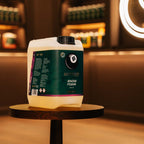 A large white plastic jug of Better Snow Foam sits on a round wooden table in a warmly lit, blurred indoor setting with shelves and a glowing circular light in the background.