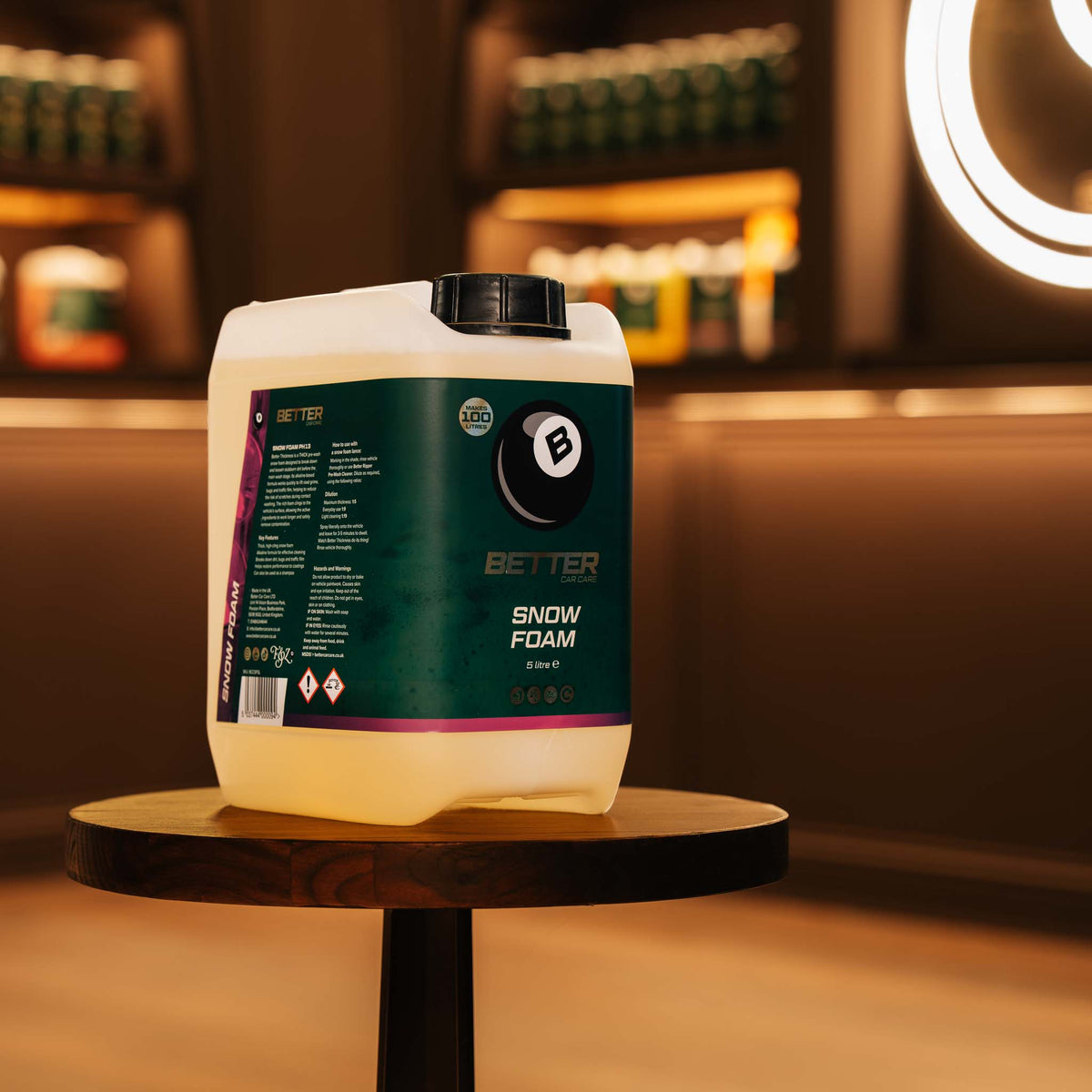 A large white plastic jug of Better Snow Foam sits on a round wooden table in a warmly lit, blurred indoor setting with shelves and a glowing circular light in the background.