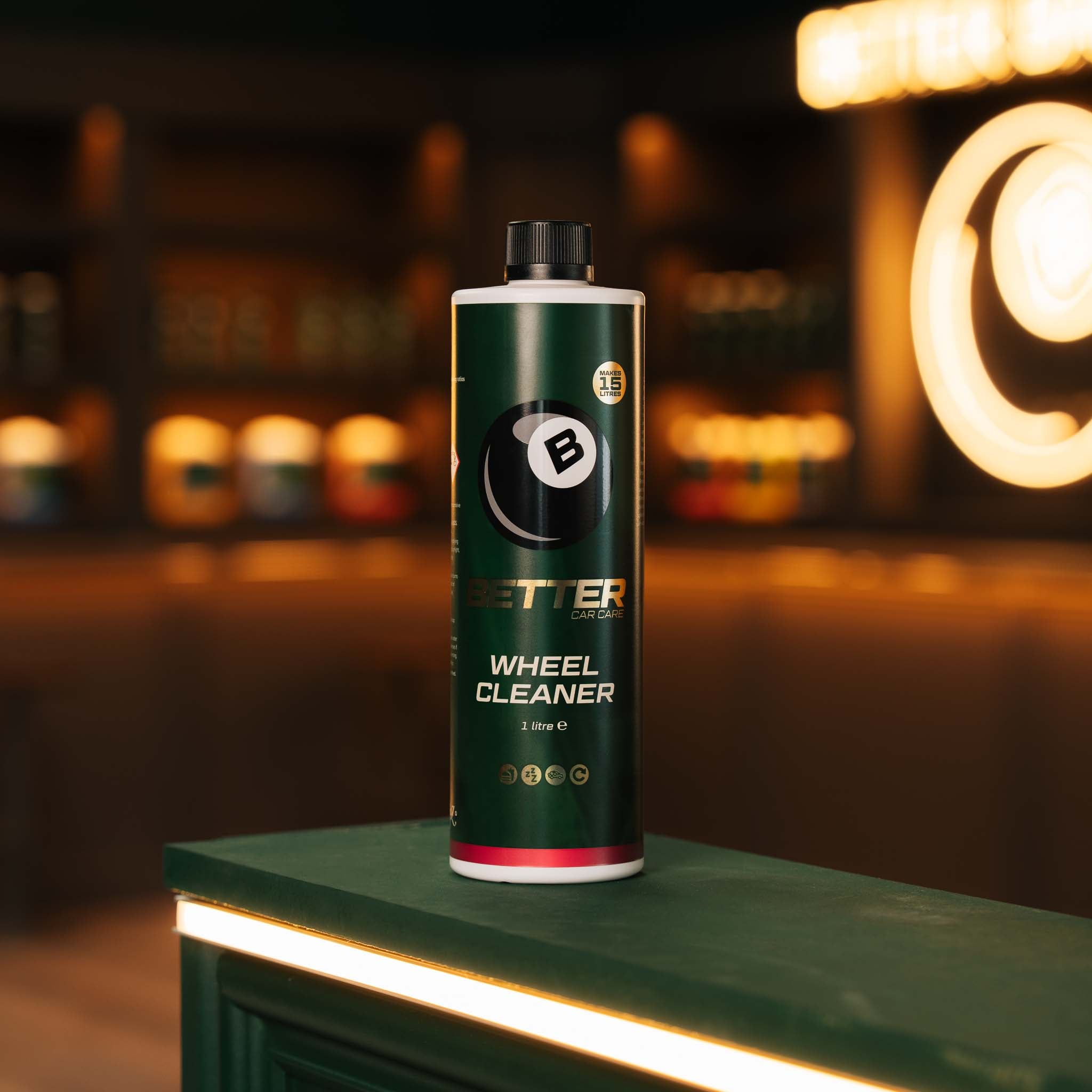 A bottle of Better Wheel Cleaner stands on a green counter in a warmly lit bar or restaurant setting with blurred lights in the background.