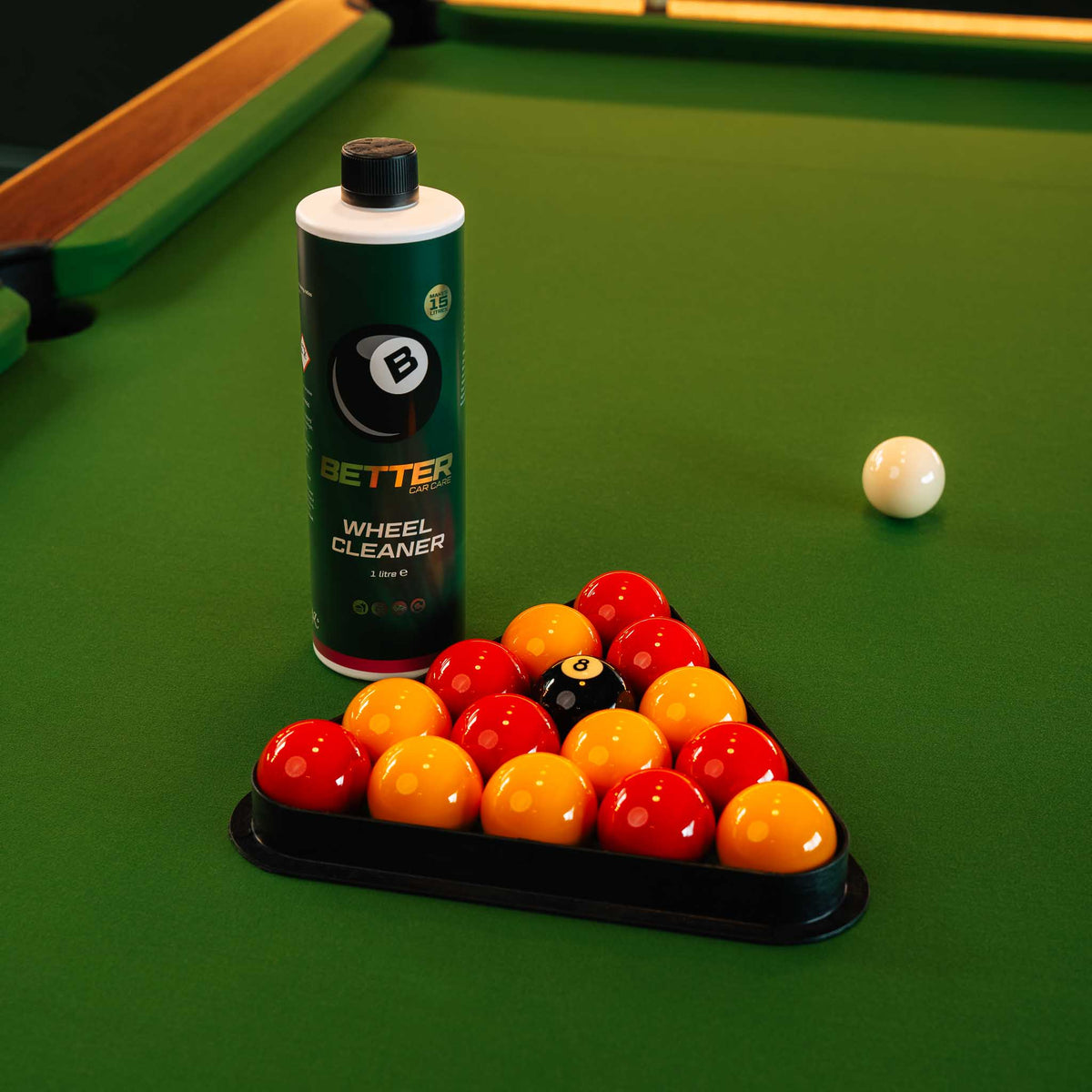 A bottle of Better Wheel Cleaner stands on a green pool table next to a black triangle rack holding colorful billiard balls, with a white cue ball nearby.